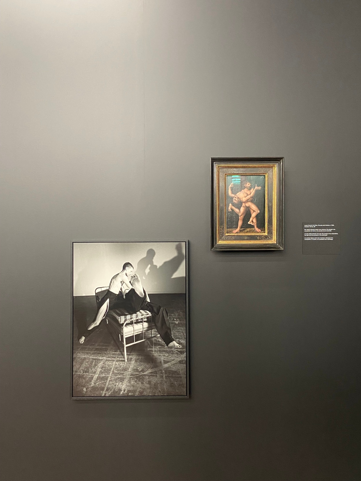 A gallery wall featuring a black and white photograph of two men fighting on a bed and a small framed classical painting of two wrestlers against a gray wall.