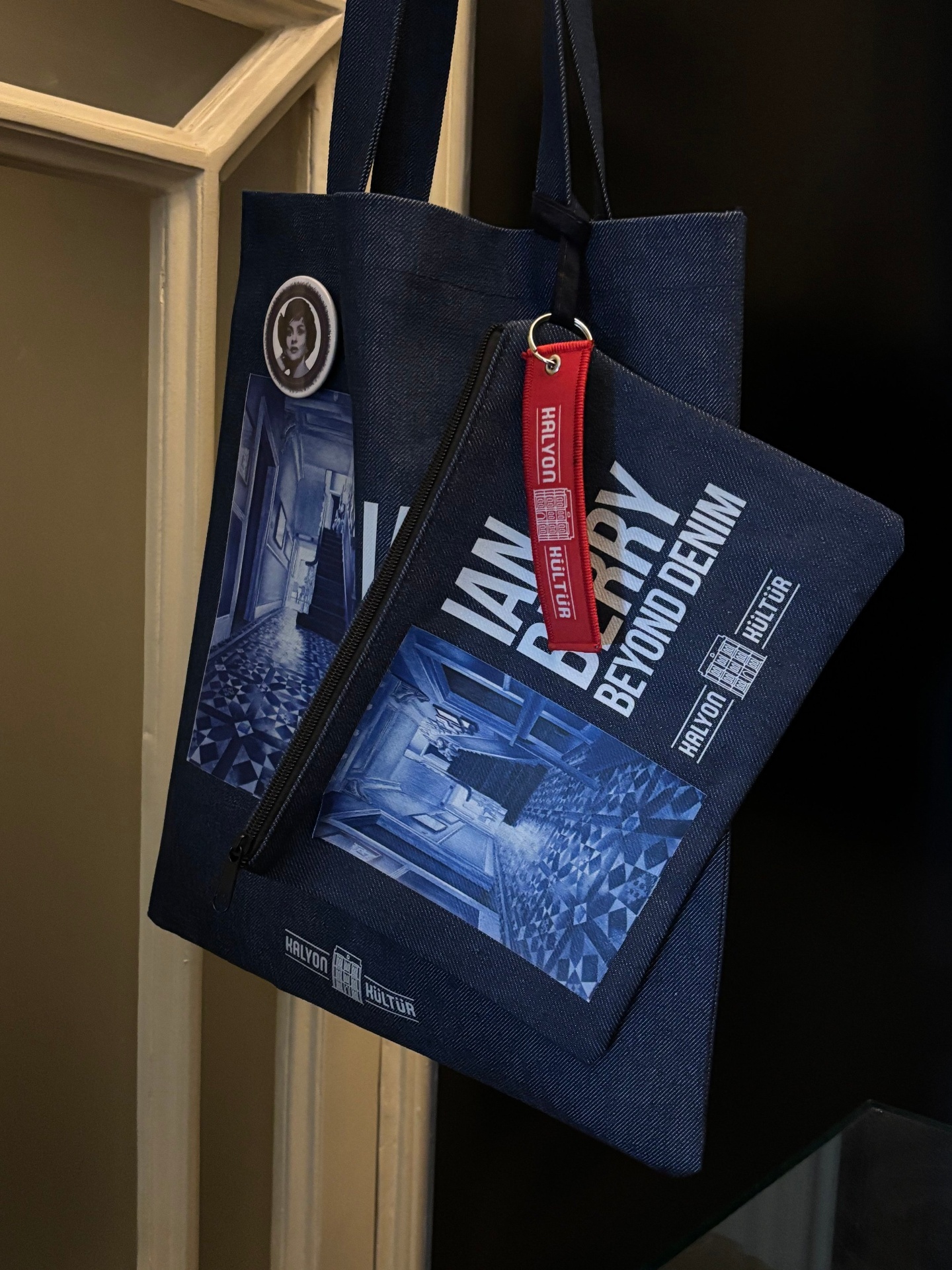 A navy blue tote bag with a printed design of an interior room and cityscape, hanging on a wooden frame. Attached is a red keychain labeled 'HULYON' and a circular badge featuring a black-and-white portrait of a woman.