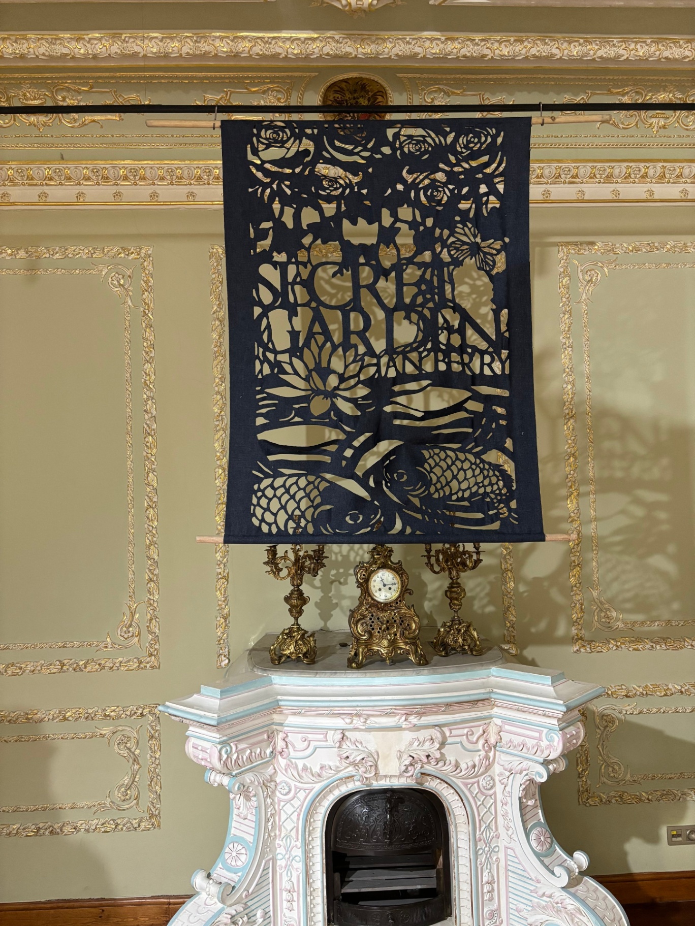 Decorative black banner with intricate floral cutouts and the text 'Sierra Garden' hanging above an ornate white fireplace with gold accents, topped with a vintage clock and two gold candelabras, in a richly decorated room.