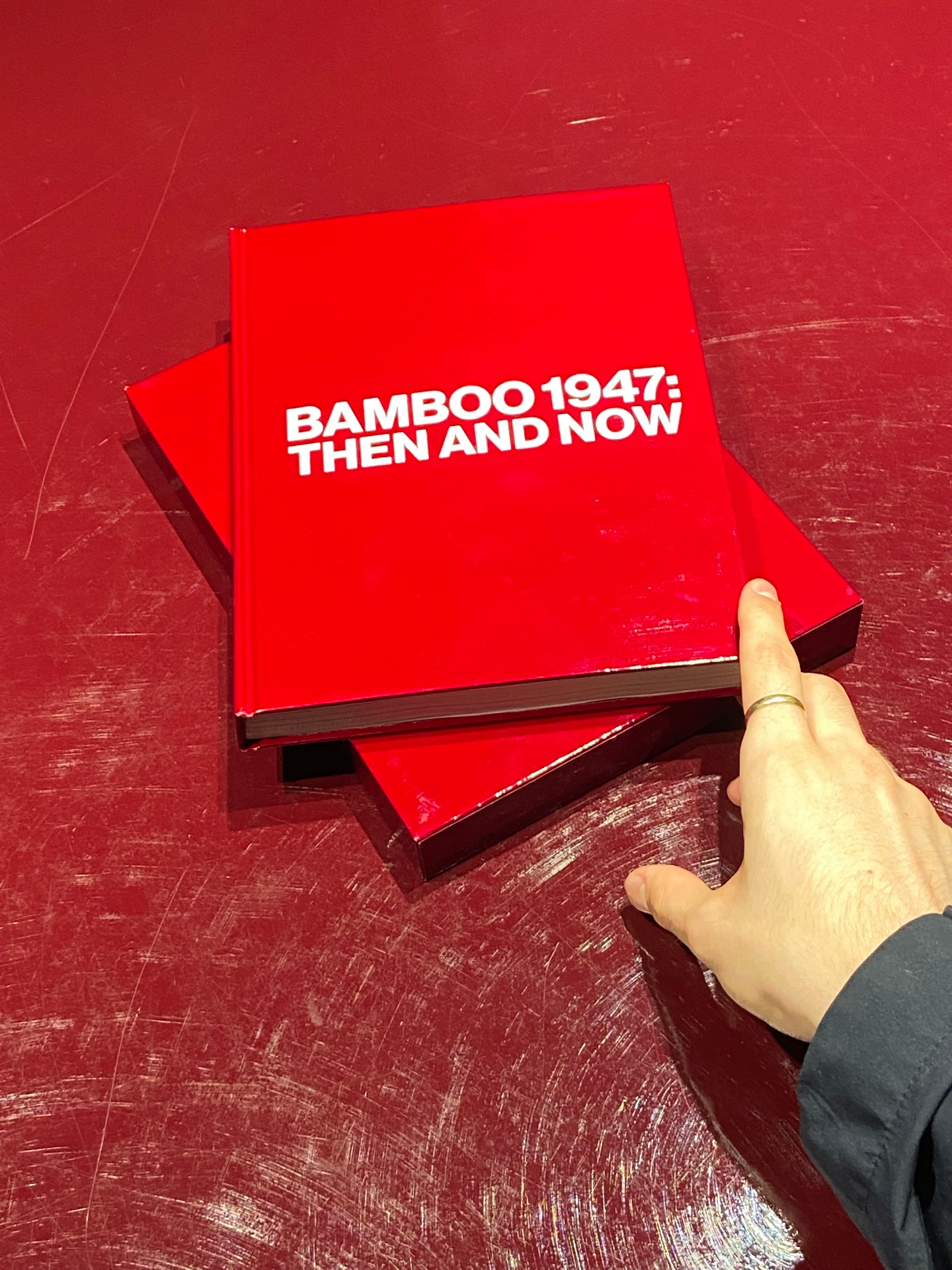 Red hardcover book titled 'BAMBOO 1947: THEN AND NOW' on a red table, with a person's finger touching the bottom right corner of the book.