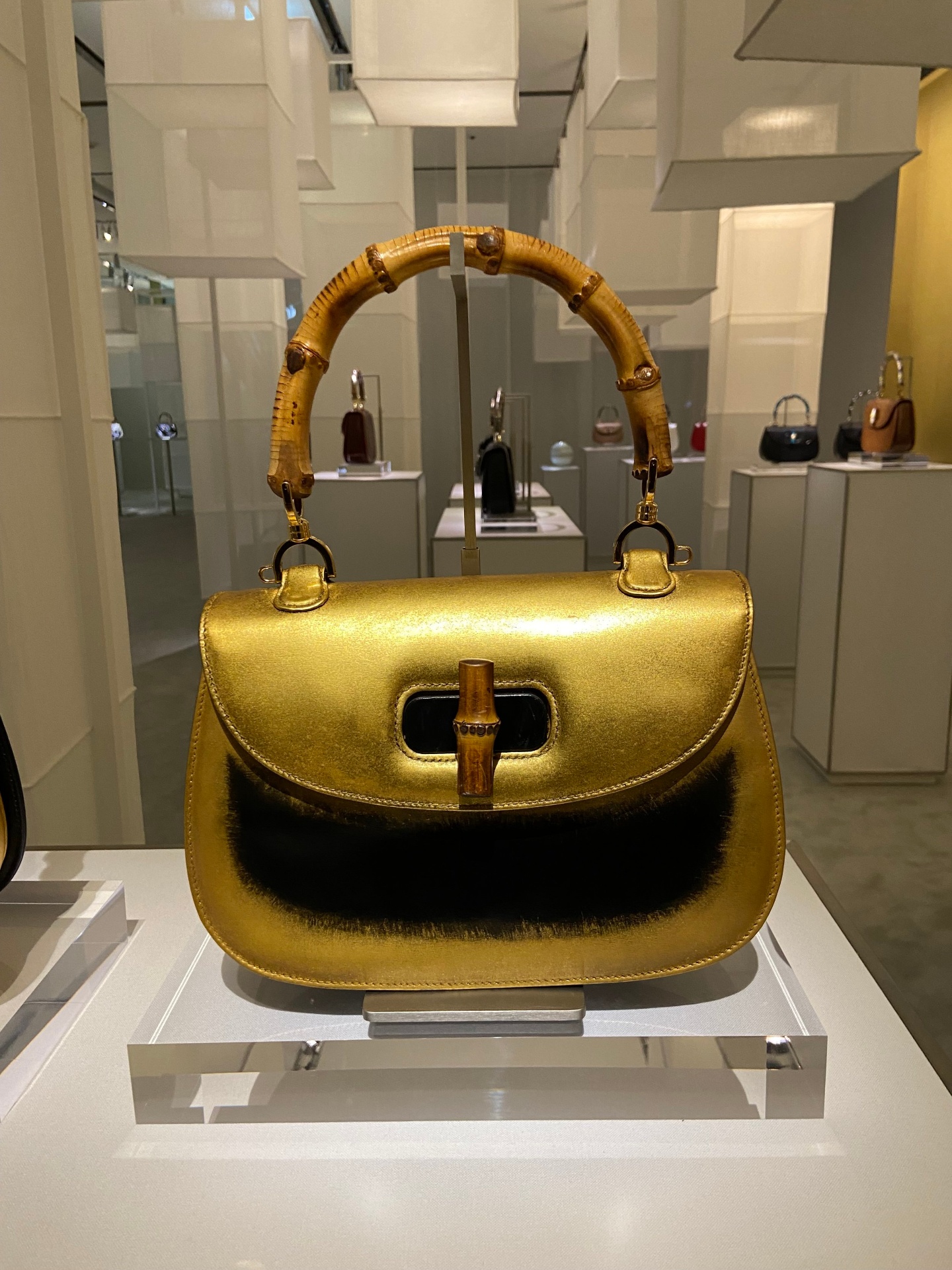 A gold handbag with bamboo handle on display in a museum or gallery setting, with other handbags in the background.
