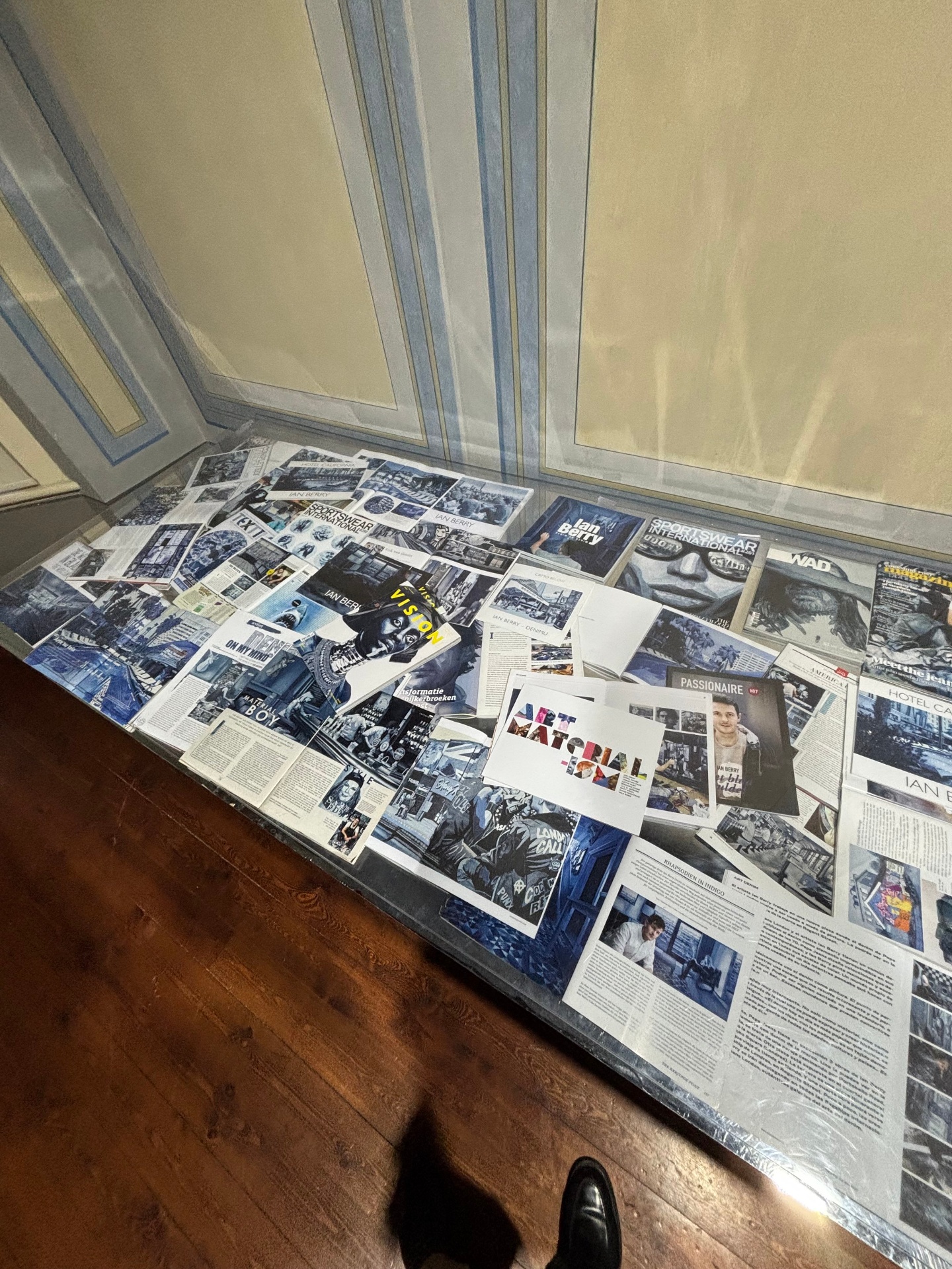 Display of magazines, newspapers, and printed materials laid out on a glass table against a wall with ornate paneling.