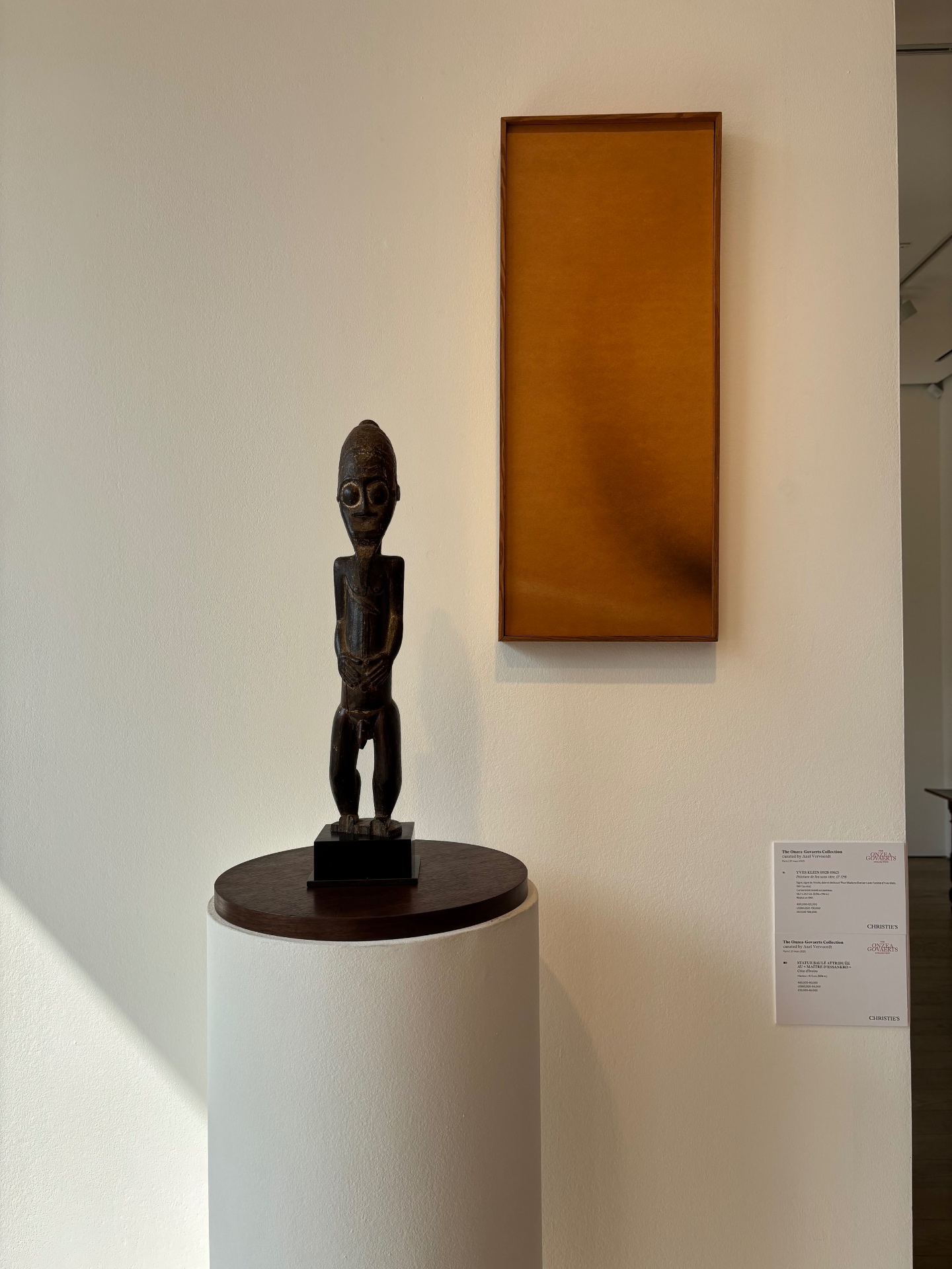 African tribal sculpture on a pedestal next to a rectangular wall art piece in an art gallery.