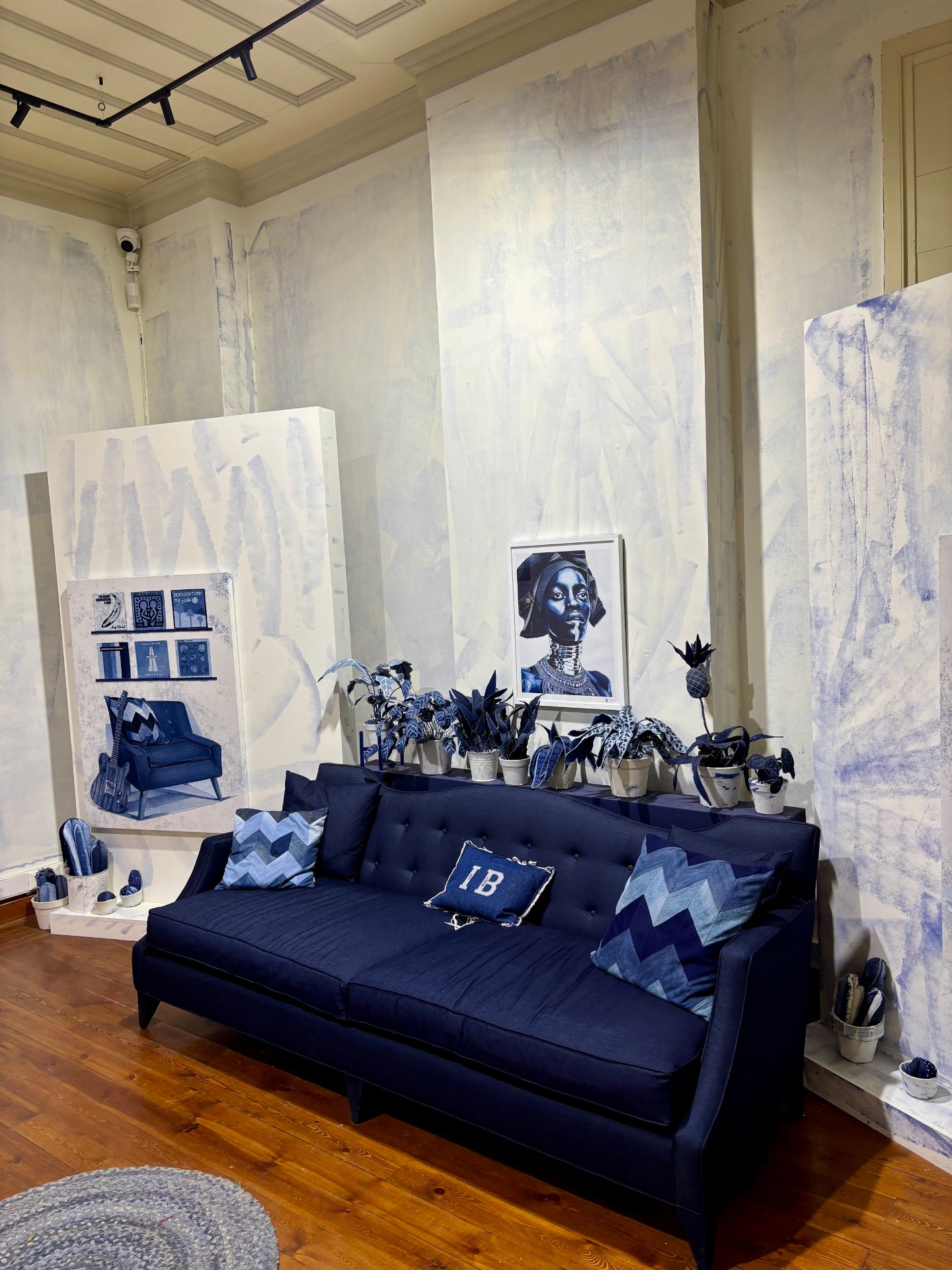Living room with a dark blue tufted sofa, decorated with patterned pillows, surrounded by potted plants on a shelf and wall, with framed artwork including a portrait and a photo of a chair and bookshelf, on a white wall with a wooden floor.