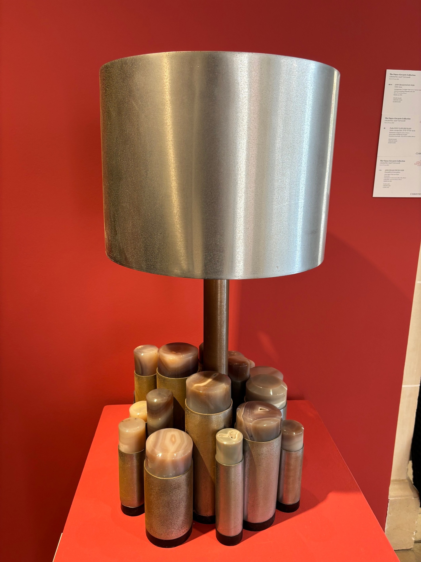 A modern table lamp with a cylindrical metallic shade and a base composed of multiple smaller cylindrical metallic objects, set against a red background.