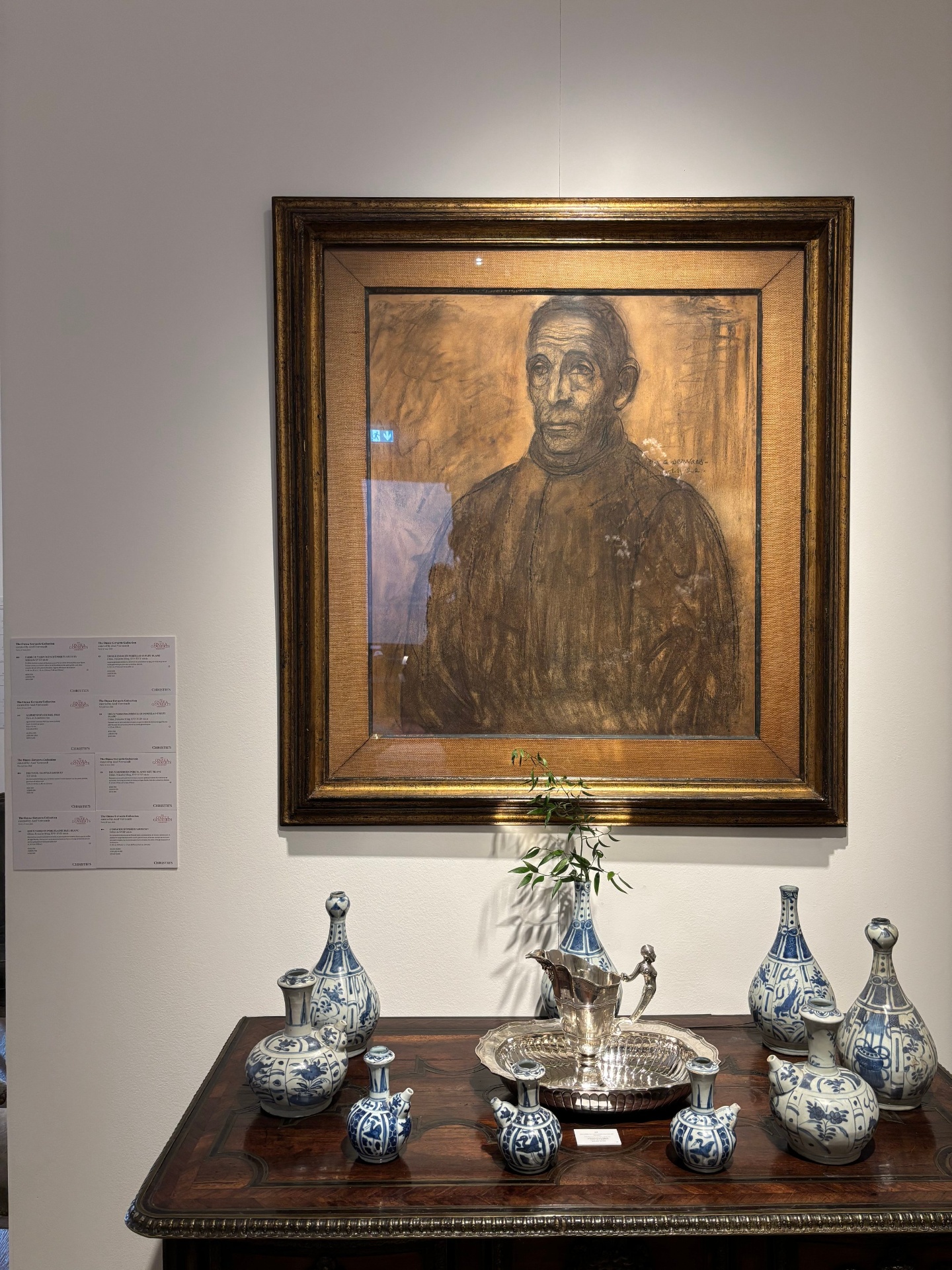 A framed portrait of a man hanging on a white wall, with a display of blue and white ceramic vases and a silver pitcher on a wooden table beneath it.