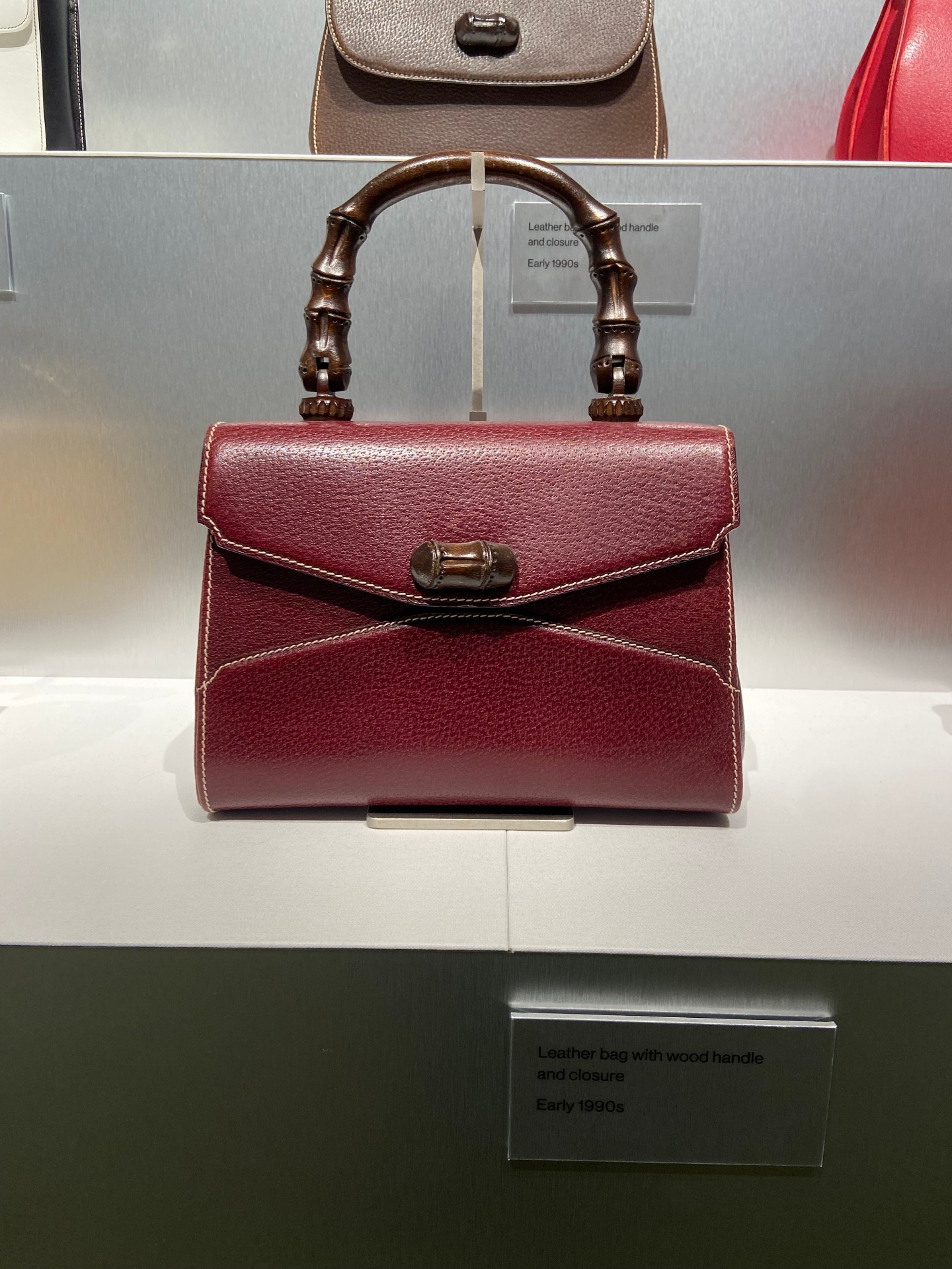 Vintage leather handbag with wood handle and closure display in a museum setting, labeled as early 1990s.