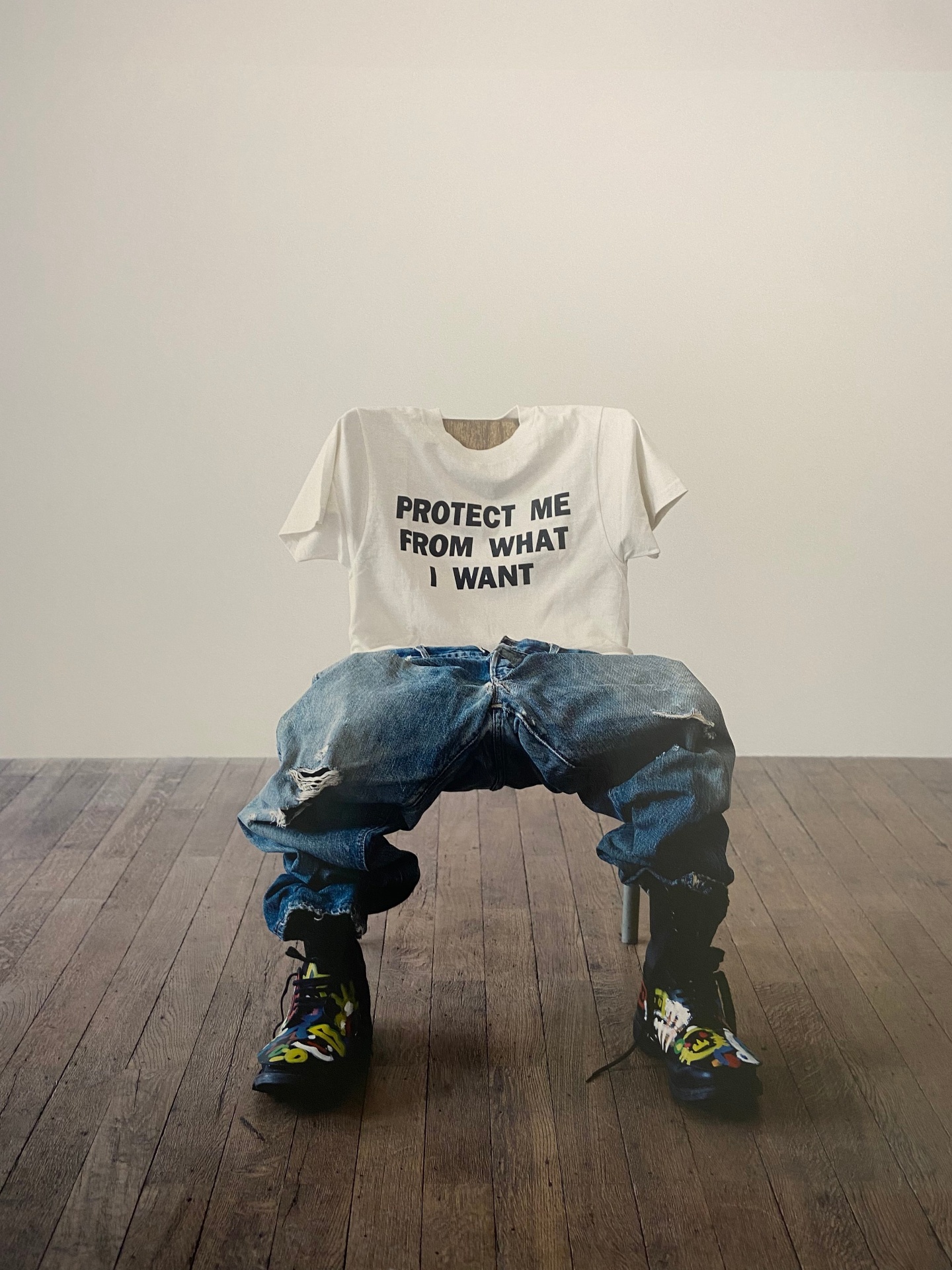 A mannequin dressed in a white T-shirt with black text, oversized ripped jeans, and colorful sneakers, seated on a stool against a plain wall.