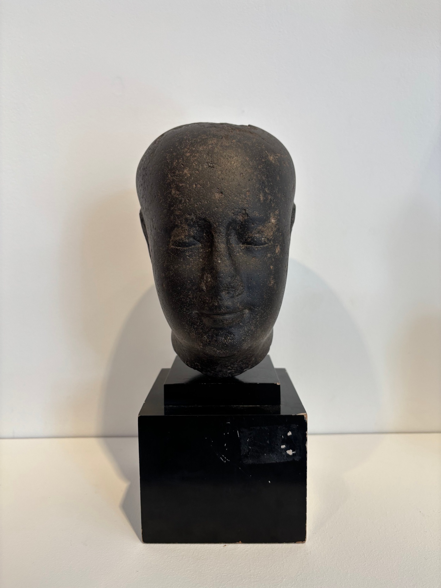 Black sculpture of a human head with closed eyes, mounted on a black rectangular pedestal.
