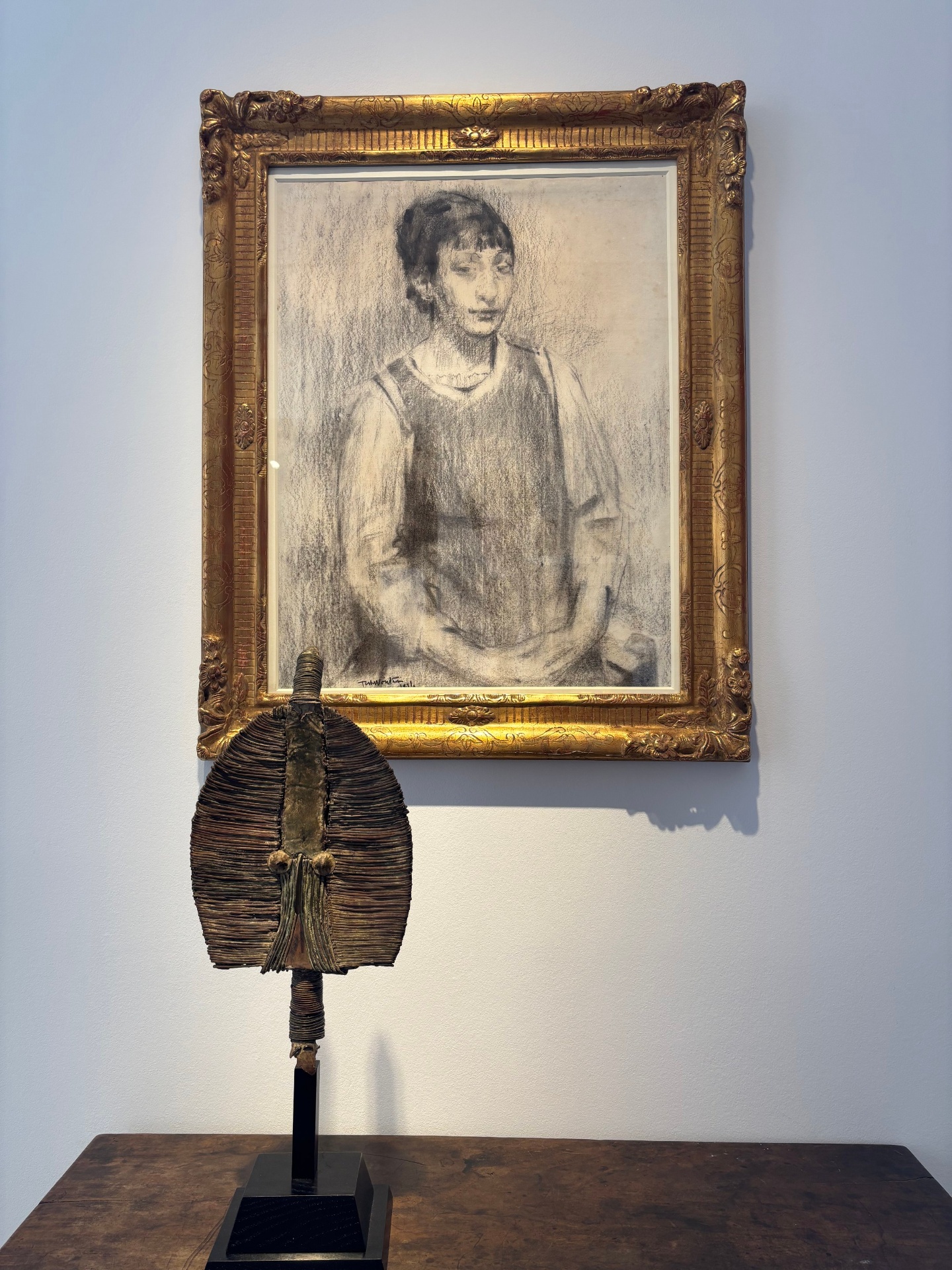 A framed pencil sketch of a young woman with short hair standing with her arms at her sides, placed on a white wall. In front of the sketch, on a wooden surface, is a sculpture resembling a stylized face or mask made of layered, textured materials.
