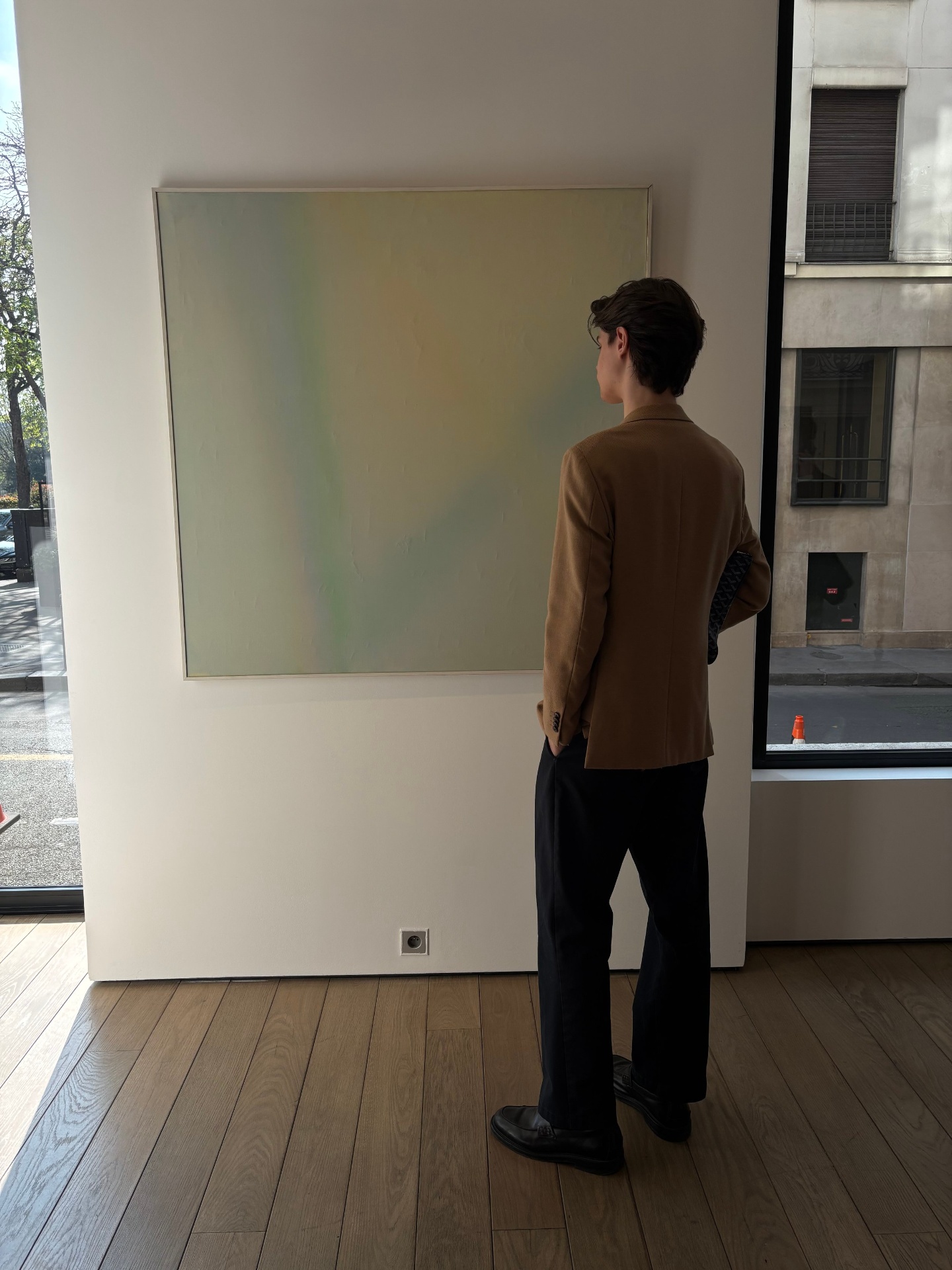 A person in a brown blazer and black pants is standing in an art gallery, facing a large abstract painting on the wall.