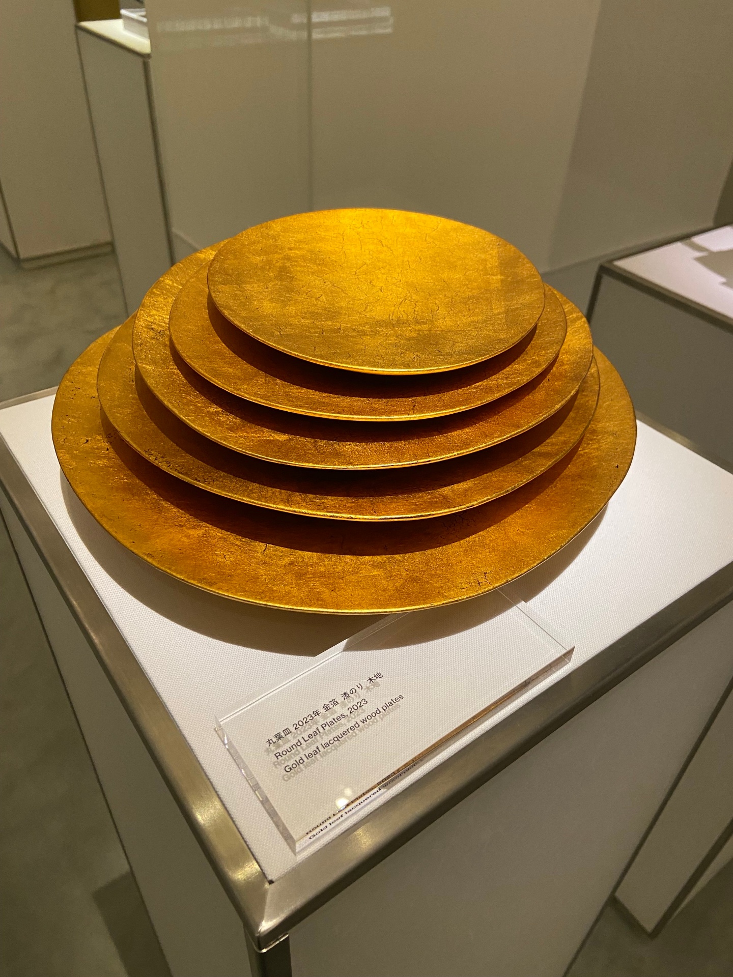 A display of five large lacquered wood plates with a gold leaf finish, shown in a museum setting on a white stand. The plates vary in size, each smaller than the one below, creating a tiered, nested appearance.