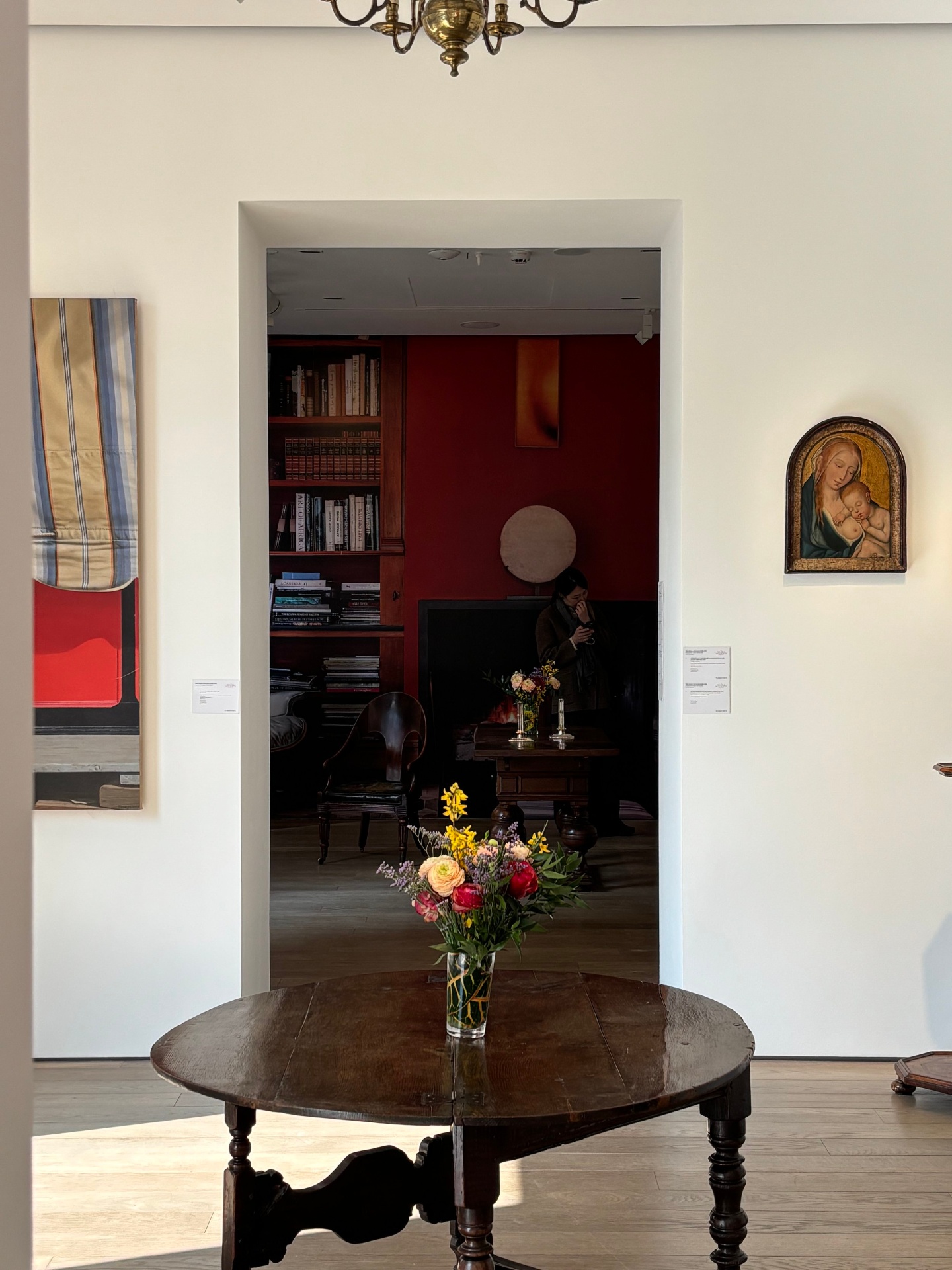 A room with a wooden table holding a vase of colorful flowers in the foreground. In the background, a doorway leads to a darker room with bookshelves, a person looking at their phone, and a small table with flowers and candles.