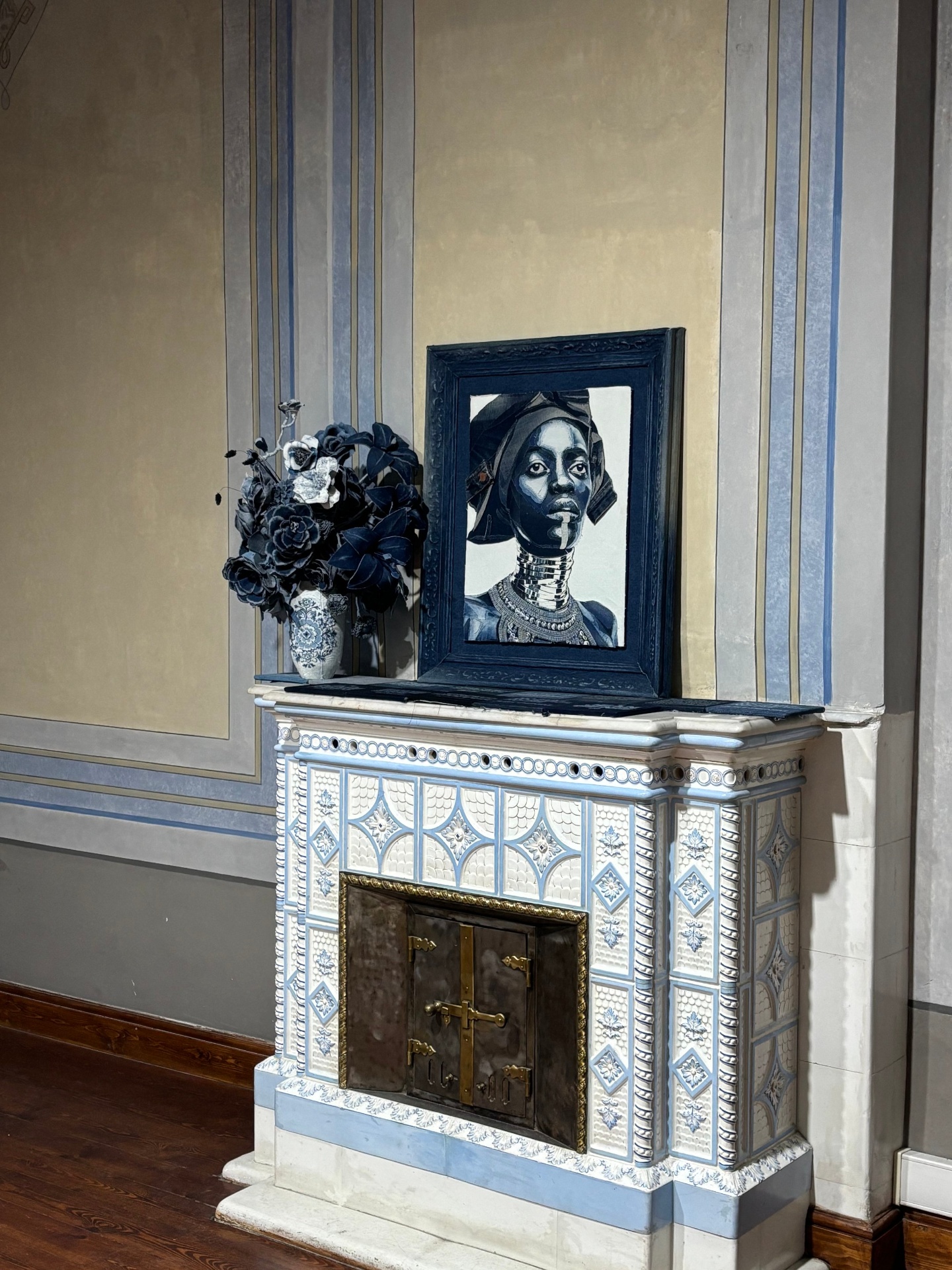 Decorative fireplace with blue and white tile design, a framed artwork of a woman with jewelry and neck rings, and a vase with dark flowers on top.