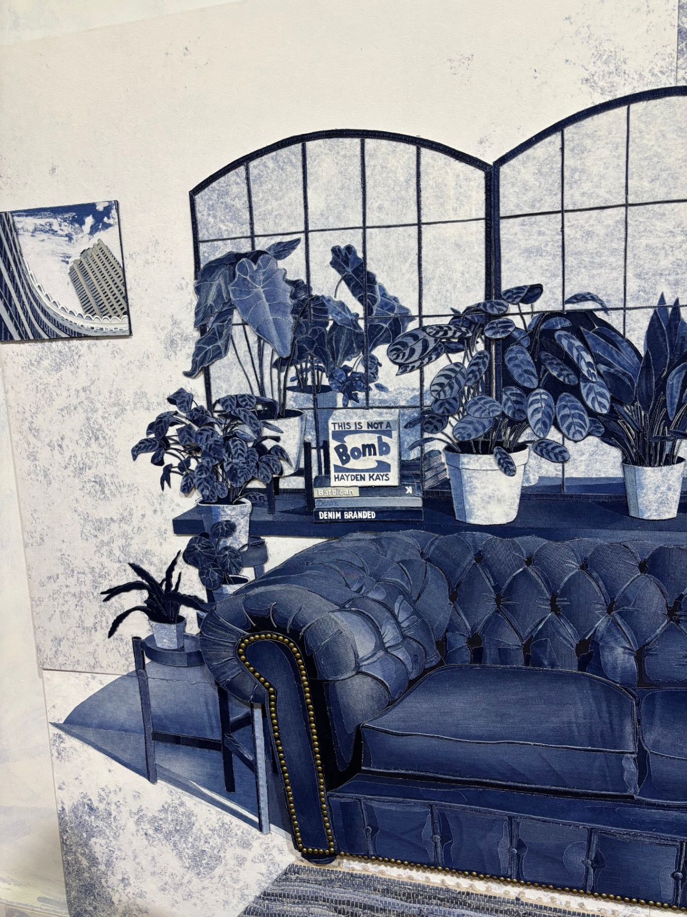 Interior of a room decorated with a dark tufted leather sofa, various potted plants on a table and windows, and a small framed sign on the table that reads 'This is NOT a Bomb'.
