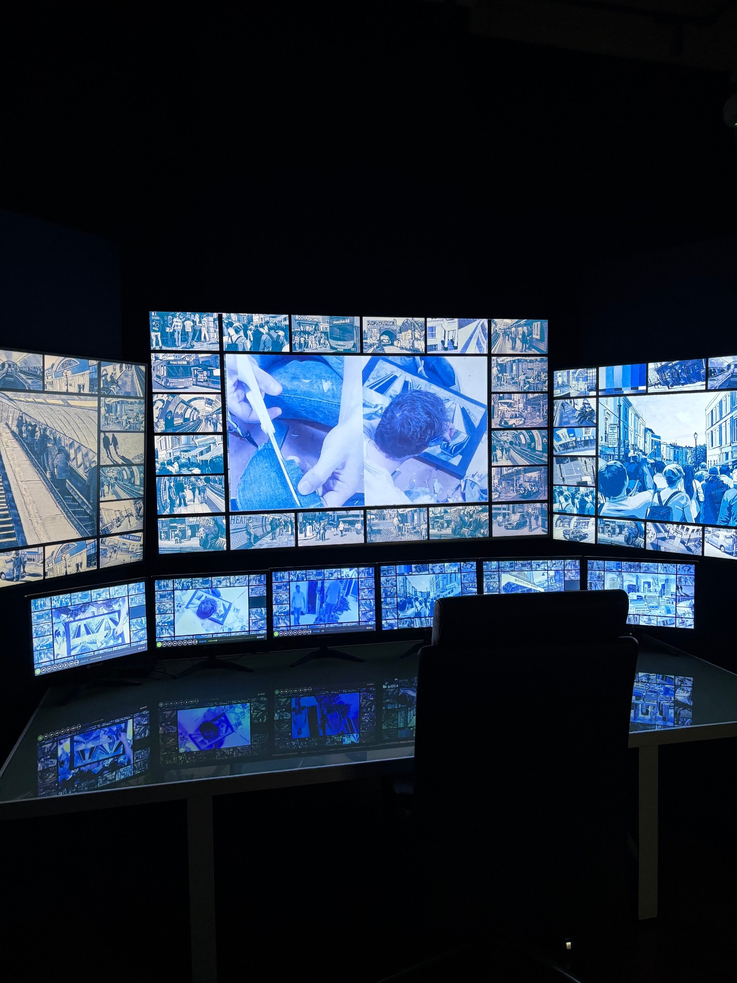 A dark room with multiple blue-tinted screens displaying various scenes, including close-ups of hands working, people in a city, and train stations, forming a large multimedia display.